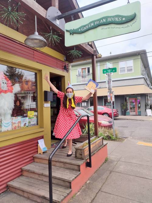 A person in a polka dot dress stands on the steps of a bookstore, smiling and holding a book, with a festive storefront behind.