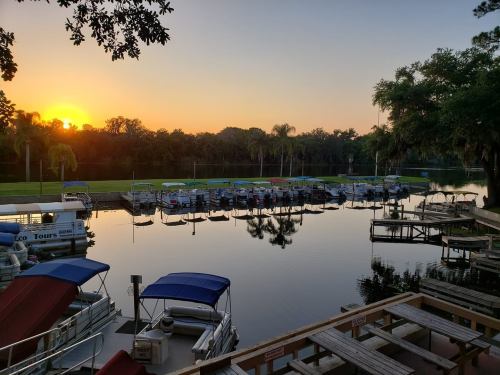 Sunset over a calm lake with boats docked, surrounded by trees and reflecting on the water's surface.