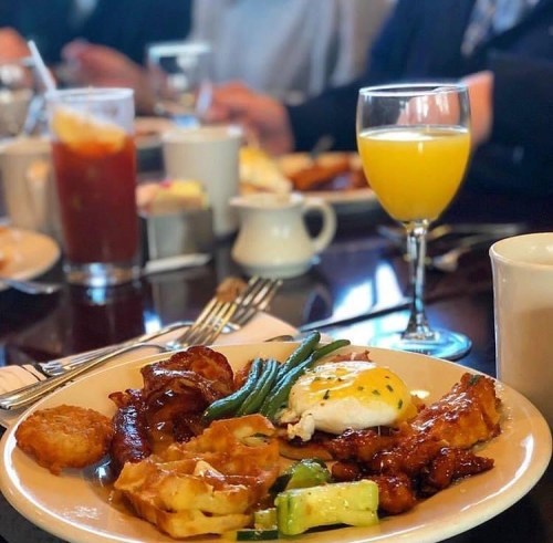 A plate of breakfast featuring waffles, eggs, green beans, and a side of potatoes, with drinks in the background.