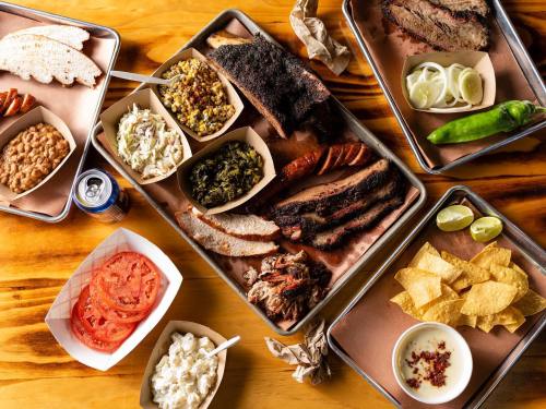 A spread of barbecue dishes on trays, including ribs, brisket, sides, and tortillas, on a wooden table.