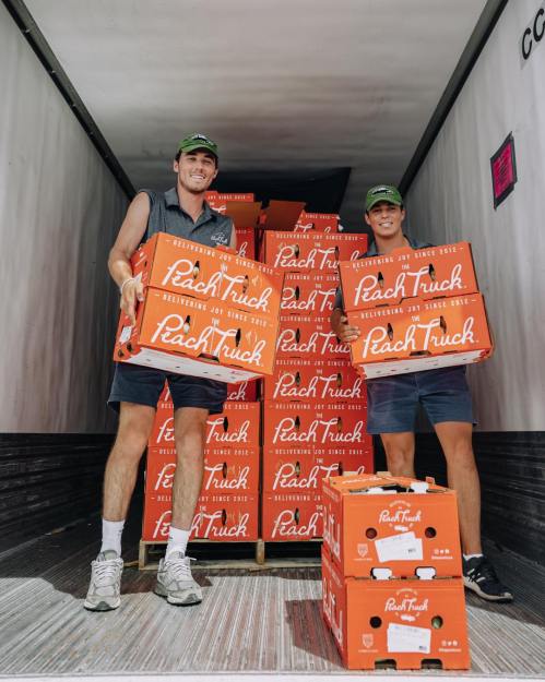 Two individuals stand in a truck filled with stacked boxes labeled "Peach Truck." They are smiling and holding boxes.