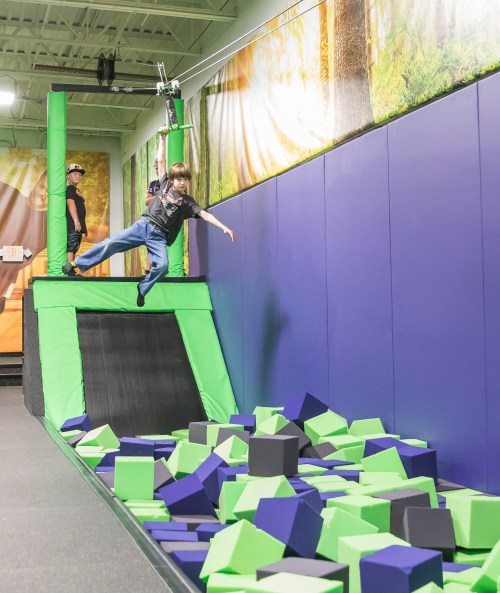 A child swings from a zip line, preparing to land on a colorful foam pit at a trampoline park.
