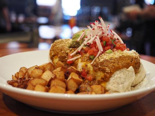 A large burrito topped with salsa, radishes, and guacamole, served with crispy potato cubes on the side.