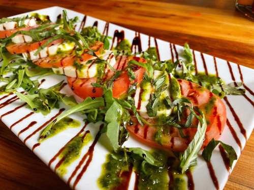 A fresh salad with sliced tomatoes, mozzarella, basil, and drizzles of balsamic and pesto on a white plate.