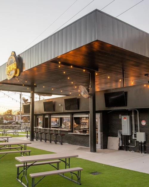 Exterior view of a modern food venue with outdoor seating, string lights, and large screens.