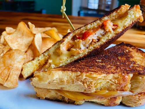 A grilled cheese sandwich with melted cheese and toppings, served with crispy potato chips on the side.