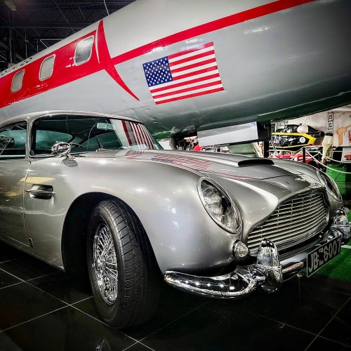 A classic silver sports car parked beside a jet, featuring a small American flag on the aircraft.