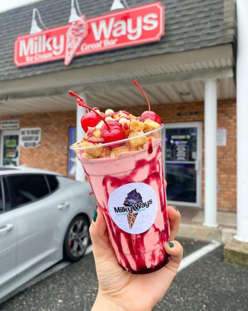 A hand holding a colorful dessert cup topped with cherries and sprinkles, in front of a Milky Ways ice cream shop.