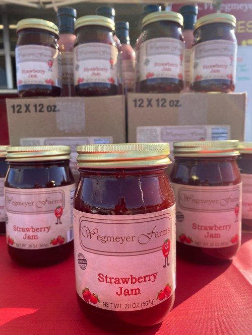 Jars of Wegmeyer Farms strawberry jam displayed in front of boxes, with a red tablecloth in the background.