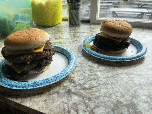 Two cheeseburgers on paper plates, each with two patties, cheese, and a top bun, sitting on a countertop.