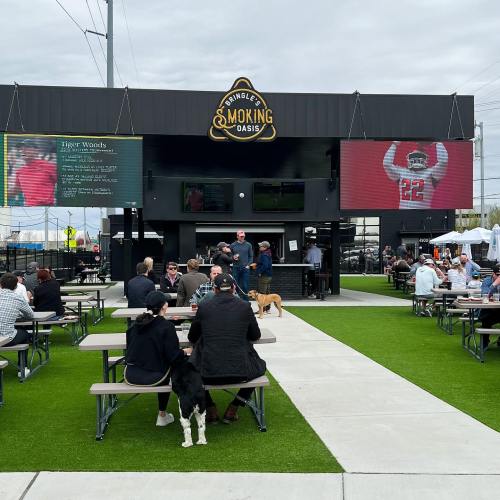 Outdoor venue with picnic tables, people socializing, and large screens displaying sports. Dogs are present.
