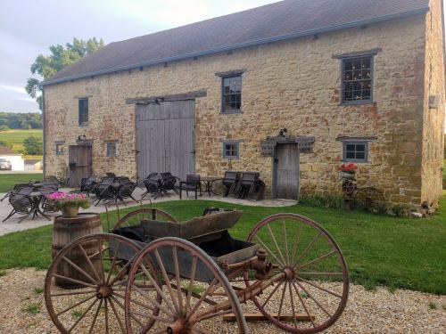 A rustic stone barn with wooden doors, surrounded by outdoor seating and a vintage wagon in the foreground.