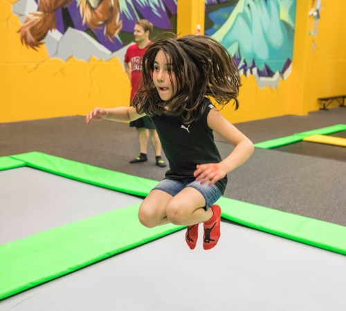A girl with long hair jumps joyfully on a trampoline in a colorful indoor trampoline park.