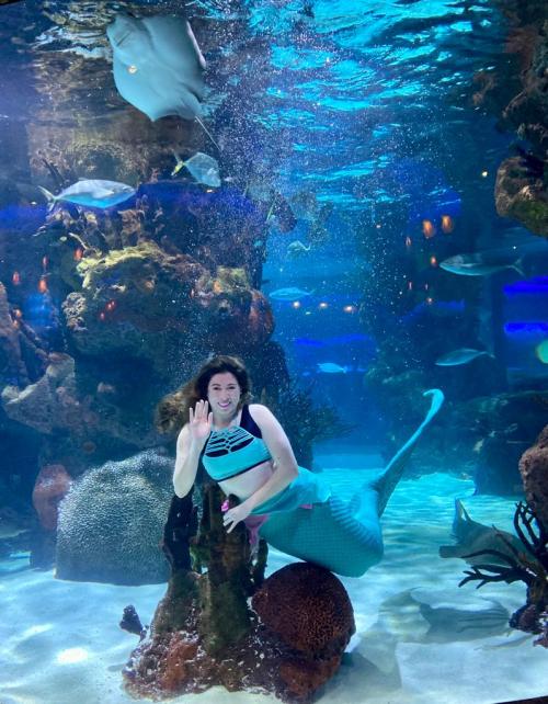 A woman waves underwater in an aquarium, surrounded by colorful fish and a stingray in a vibrant marine environment.