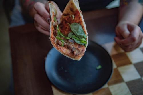 A person holds a slice of pizza topped with greens over a black plate on a wooden table.