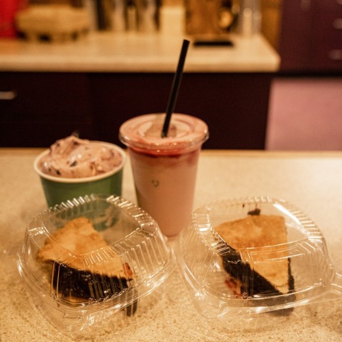 Two slices of pie in clear containers, a cup of ice cream, and a smoothie with a straw on a countertop.