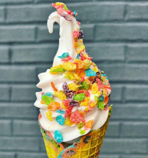 A colorful ice cream cone topped with swirled soft serve and vibrant cereal sprinkles against a textured gray background.