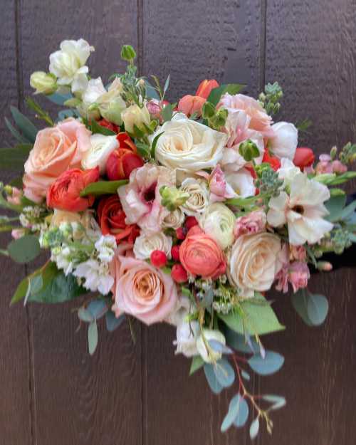 A vibrant bouquet of roses, peonies, and greenery in soft peach, pink, and white tones against a wooden background.