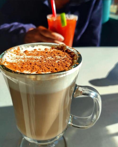 A close-up of a latte topped with cinnamon, with a blurred background of a person holding a drink.