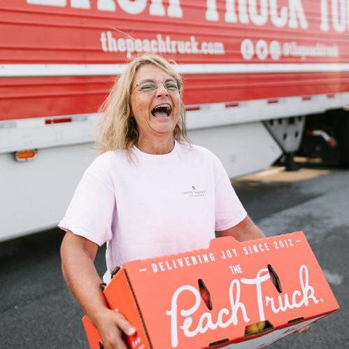 A smiling woman in a pink shirt holds a box of peaches in front of a red truck with "The Peach Truck" logo.