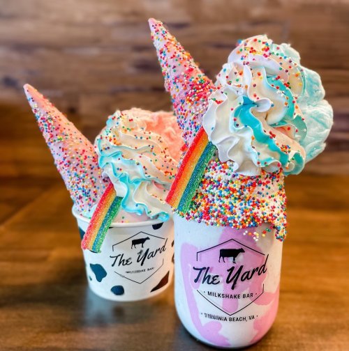 Two colorful milkshakes topped with whipped cream, cotton candy, sprinkles, and rainbow cones, served in branded cups.