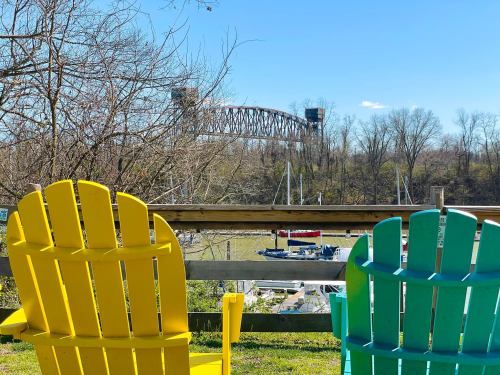 Two colorful adirondack chairs in the foreground, overlooking a river with a bridge and boats in the background.