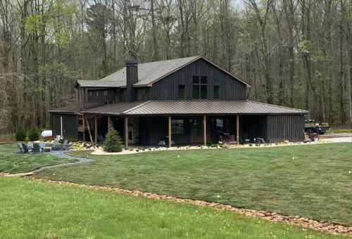 A modern two-story house with a metal roof, surrounded by trees and a grassy yard, featuring a patio area with seating.