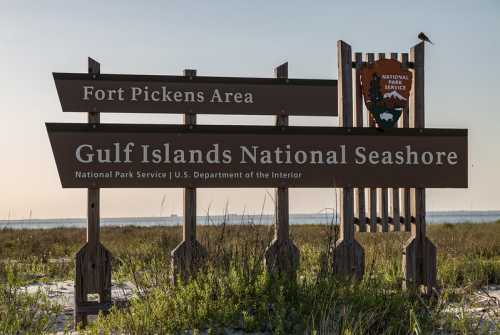 Sign for Gulf Islands National Seashore, Fort Pickens Area, with a bird perched on top and a coastal view in the background.