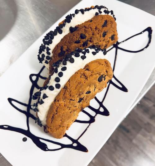 Two cookie sandwiches filled with cream and topped with chocolate chips, drizzled with chocolate sauce on a white plate.