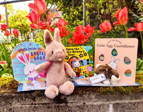 A plush bunny sits among colorful Easter-themed books and vibrant tulips in the background.