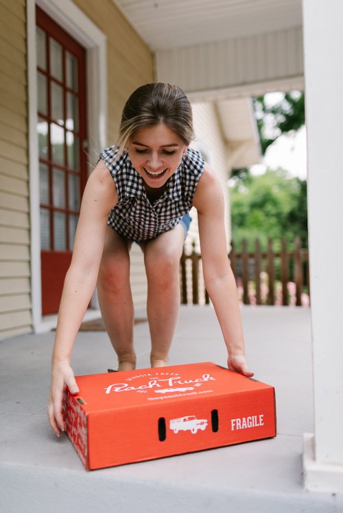 A woman in a checkered top smiles as she lifts a red box labeled "FRAGILE" on her porch.
