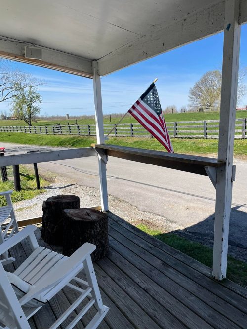 A porch with rocking chairs and a small American flag, overlooking a grassy field and a quiet road.