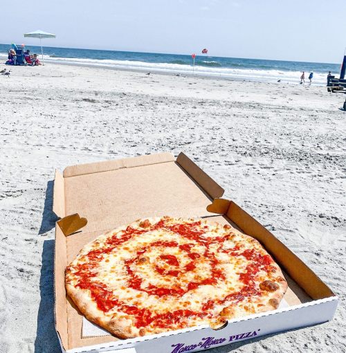 A large pizza in an open box on a sandy beach, with the ocean and beachgoers in the background.