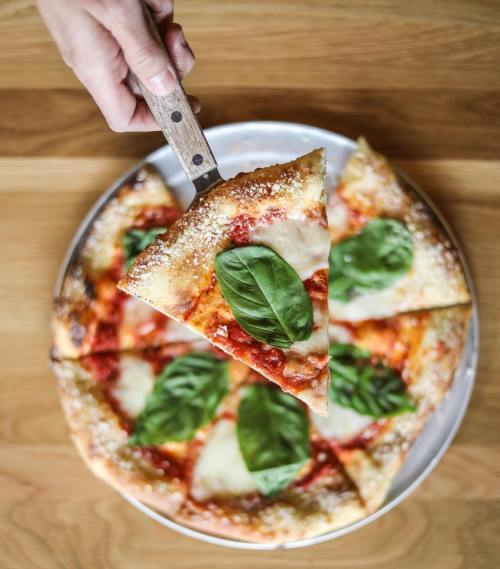 A hand lifts a slice of pizza topped with basil from a round pizza on a wooden table.