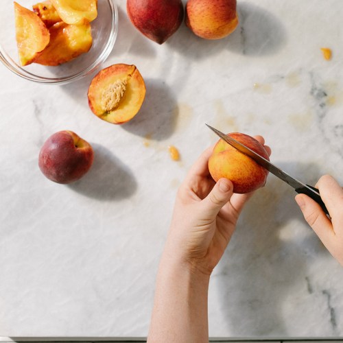 A hand holding a peach while cutting it, with sliced peaches and a bowl in the background on a marble surface.