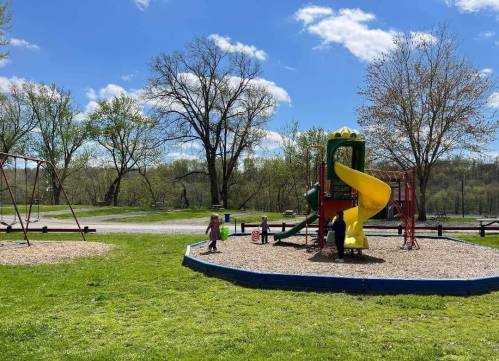 Children playing on a colorful playground with a slide, surrounded by green grass and trees under a blue sky.
