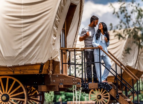 A couple stands on the steps of a cozy wagon, holding mugs and smiling at each other in a scenic outdoor setting.