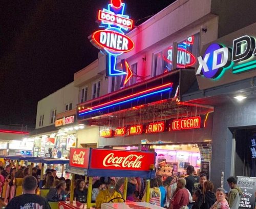 A vibrant diner with neon lights and a Coca-Cola cart, bustling with people on a lively boardwalk at night.