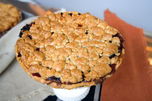 A freshly baked pie with a golden, crumbly topping and a dark fruit filling, displayed on a white pedestal.