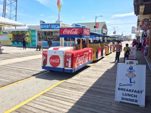 A colorful Coca-Cola train on a boardwalk, with food stands and people enjoying the sunny day.
