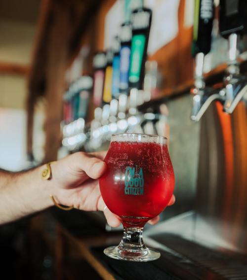 A hand holds a glass of red beverage in front of a row of colorful beer taps.