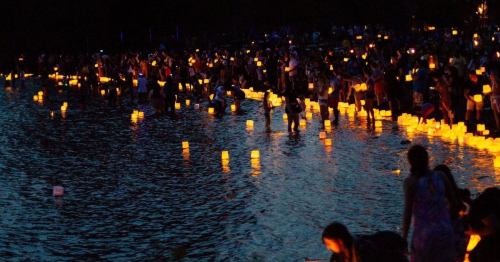 A crowd gathers by a water's edge, releasing glowing lanterns into the dark, reflecting on the surface.