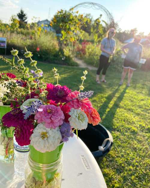 A close-up of colorful flowers in jars on a table, with two people in the background enjoying a garden setting.