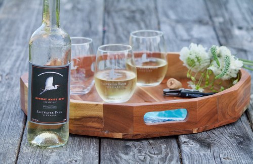 A wooden tray holds a bottle of white wine, four glasses of wine, and a small bouquet of flowers on a rustic table.