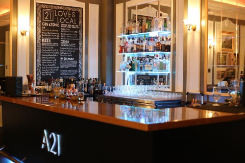 A stylish bar with a wooden counter, shelves of various spirits, and a chalkboard menu featuring drinks.