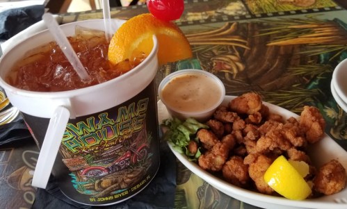 A colorful drink with a cherry and orange slice beside a plate of fried seafood and dipping sauce on a vibrant table.