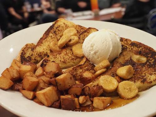 A plate of French toast topped with bananas and a scoop of ice cream, served with diced, roasted potatoes.