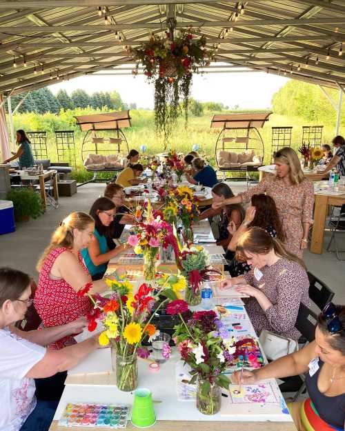 A group of people creating art with flowers on tables, surrounded by greenery in a spacious outdoor setting.