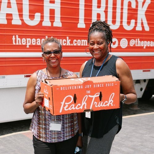 Two women smile while holding a box from The Peach Truck in front of a red truck with the company logo.