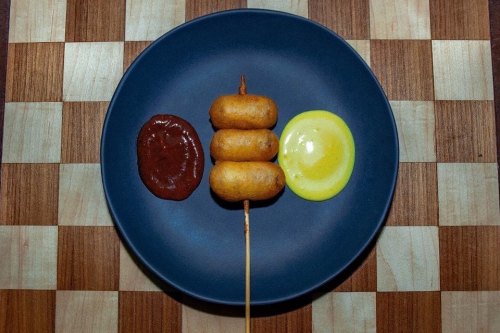 A plate with three corn dogs on a skewer, accompanied by ketchup and mustard on a wooden checkerboard background.